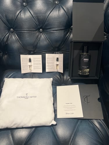 Reviewer's Smoke & Mirrors Eau de Parfum bottle in its open box, displayed with samples and a branded pillow on a leather seat.