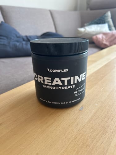 Customer's black tub of Complex Creatine Monohydrate sitting on a wooden table in a living room setting.