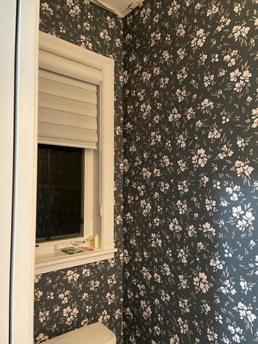 Customer's bathroom showing the dark floral Alina Wallpaper installed in a corner around a white-trimmed window.
