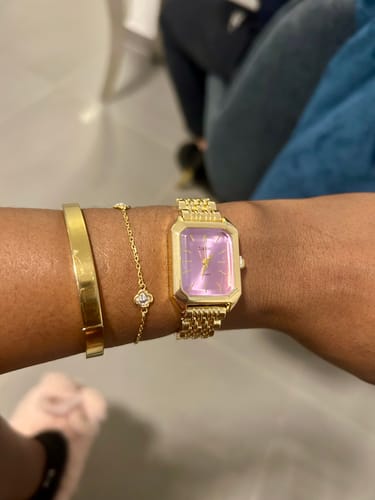 Reviewer showing their CLAIRE DE MAINS watch. The gold timepiece has a rectangular purple dial and is worn with two bracelets.