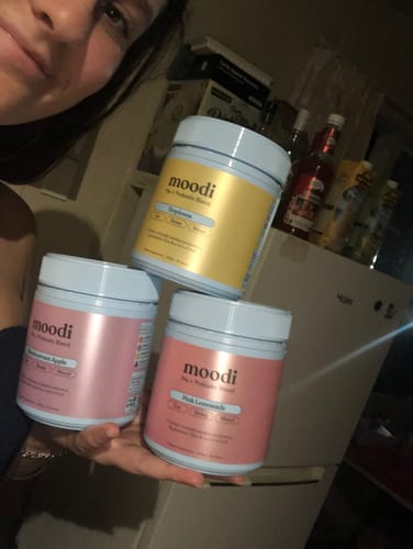 Reviewer holding three different jars of Moodi supplement powder: Tropicana, Raspberry Apple, and Pink Lemonade.
