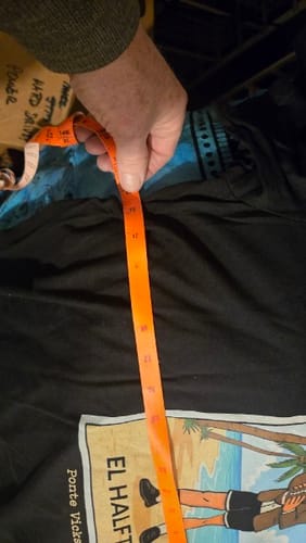 Customer using a tape measure to show the chest measurement of the black El Halftime OG T-Shirt.