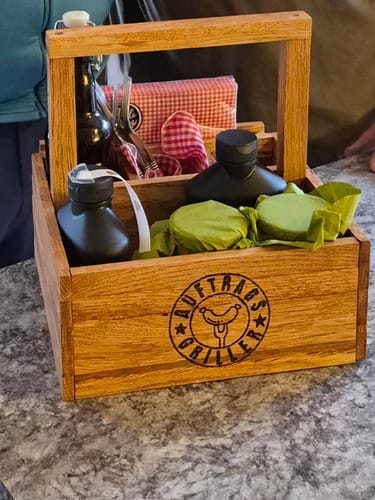 Kunde zeigt einen selbstgebauten Grillbutler aus Holz, gefüllt mit Flaschen und Grillzubehör auf einer Theke.