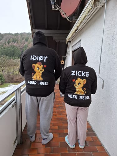 Zwei Rezensenten von hinten, die passende schwarze Premium Unisex Hoodies mit den Aufdrucken 'Idiot' und 'Zicke' auf einem Balkon tragen.