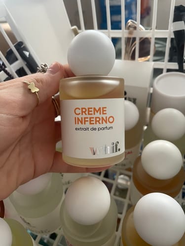 Customer holding a bottle of Creme Inferno perfume, showing its white label and round white cap in front of a fragrance collection.