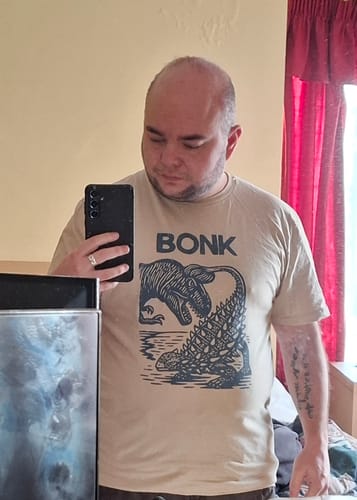 Customer taking a mirror selfie wearing the sand-colored Late Cretaceous Bonk T-Shirt, showing the fit and graphic.