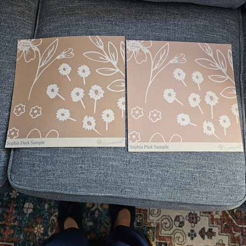 Customer compares two floral Sophia Wallpaper samples, one dark tan and one light pink, on a couch.