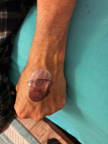 Customer's hand with a clear custom photo face temporary tattoo of a man with glasses.