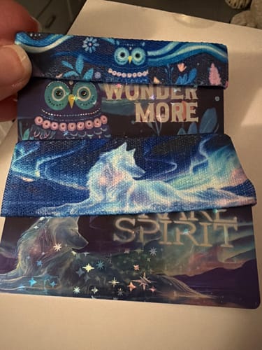 Customer holding two bracelets and cards from the Aurora Aura Collection: the 'Wonder More' owl and 'Rare Spirit' wolf designs.