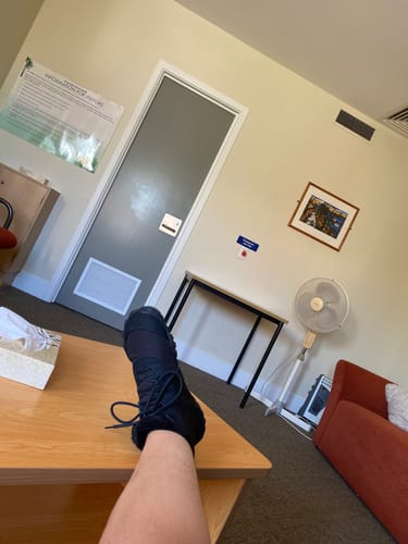 Customer's foot wearing a black Will II Winter Barefoot Shoe, resting on a wooden table indoors.