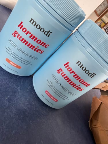 Customer's two light blue jars of Moodi Hormone Gummies, one Peach and one Strawberry flavor, on a dark surface.