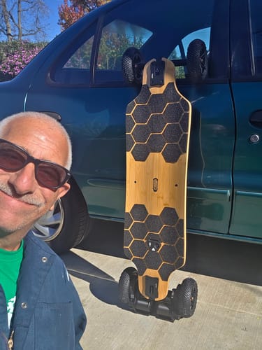 Reviewer taking a selfie with their new MEEPO ROVER electric skateboard standing upright next to a car outdoors.