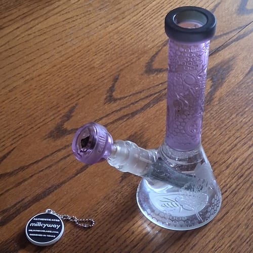 Reviewer's purple Apiary Mini Beaker Bong with etched bee designs on the glass base, sitting on a table.