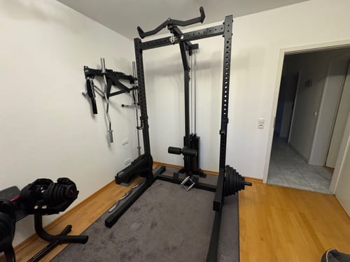 Das schwarze R7 Rider Squat Rack eines Kunden, aufgebaut in einem Zimmer mit Latzug und Hanteln.