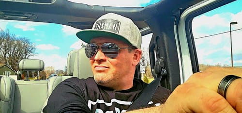 Reviewer wearing a gray Indiana Blackout Plate Hat with a flat bill and 'THXMAN' text while driving.