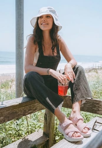 Reviewer wearing pink Haloproduct sandals while sitting on a wooden railing at the beach.