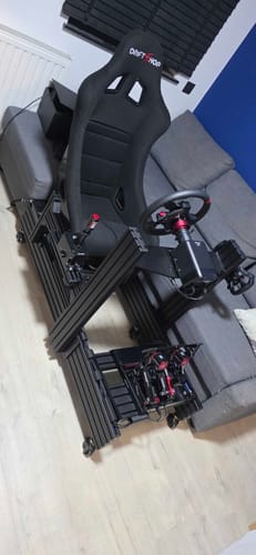 Customer's black PRIME Cockpit fully assembled with a racing seat and wheel, positioned next to a sofa.