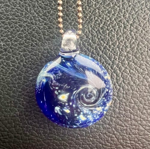 Close-up of a customer's blue Nightswirl Pendant with a starry swirl design on a black surface.