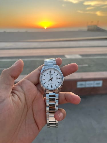 Un cliente sostiene su reloj analógico Casio con esfera blanca frente a un atardecer.