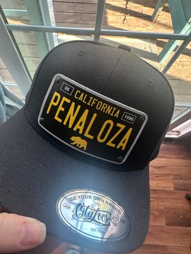 Customer holds a black California 60's Plate Hat with a custom license plate patch that reads 'PENALOZA'.