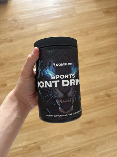 Customer's hand holding the black tub of Complex Sports Iont Drink against a wood floor.