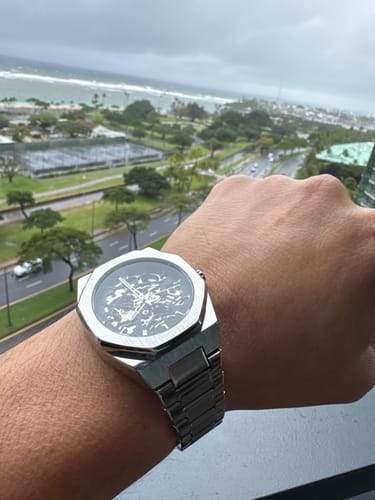 Reviewer's silver Lucky Fours® Spine watch with a skeleton dial on their wrist, overlooking a coastal city.