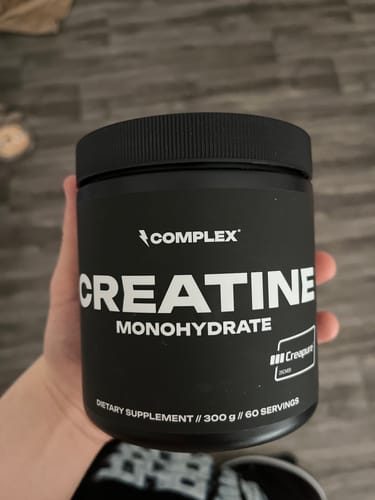 A customer's hand holding a black tub of Complex Creatine Monohydrate supplement against a blurred background.