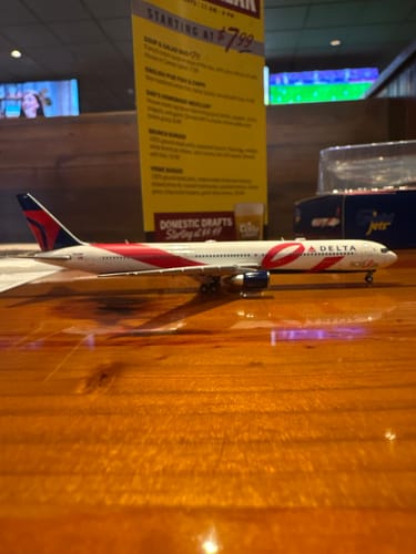 Customer's GEMINI DELTA 767-400ER model airplane with breast cancer awareness livery, displayed on a wooden table in a restaurant.