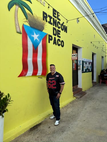 Customer wearing a black Puerto Rico Heritage Baseball Jersey with red number 62, standing by a yellow wall with a PR flag.