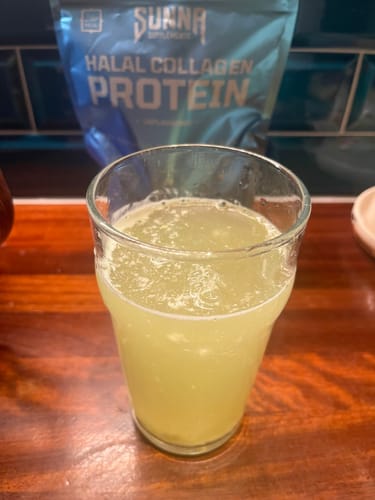 Customer's glass of a prepared yellow drink, with a pouch of Halal Collagen Protein in the background on a counter.