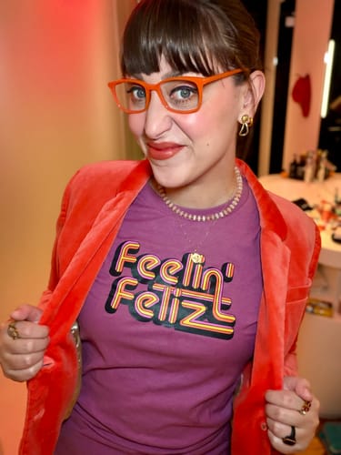 Reviewer opens an orange jacket to show the mulberry Unisex Feelin Feliz Tee with its colorful graphic.