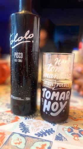 Un cliente muestra un vaso del juego 'Hoy me lanzo' con una bebida oscura. El vaso tiene la frase 'No tomes mañana lo que puedes tomar hoy'.