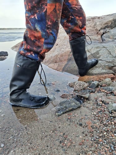 Asiakkaan mustat Vaistoa Katve 2 -paljasjalkakumisaappaat märällä ja kivikkoisella rannalla.