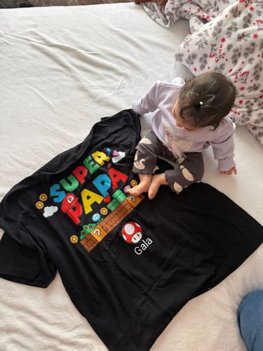 Ein Kunde zeigt das personalisierte schwarze T-Shirt 'Super Papá', das neben einem Baby auf einem Bett liegt.