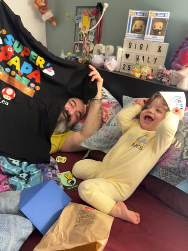 Ein Kunde hält spielerisch das schwarze 'Super Papà'-T-Shirt hoch, während er mit einem Kind im Bett liegt.