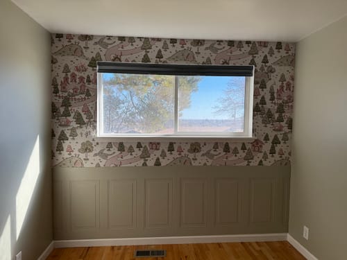 Customer's nursery with Adelina Wallpaper on the upper half of a wall, above tan wainscoting and surrounding a window.