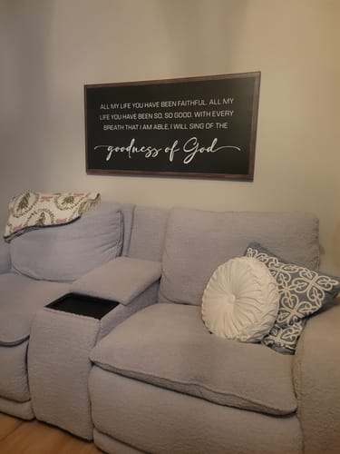 Reviewer's dark-framed Goodness of God Faith Sign hanging on the wall above a grey couch.