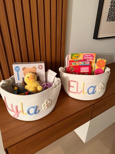 Customer's two personalized woven baskets, named Lyla and Ella, filled with books and toys on a wood surface.