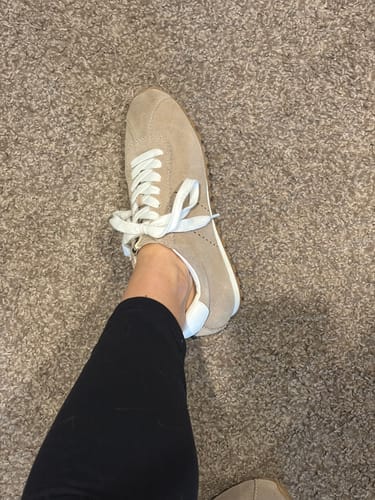 Close-up side view of a reviewer's foot in the beige suede PRISCILLA sneaker on a carpeted floor.