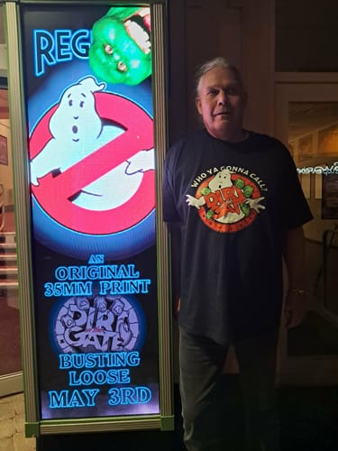 Customer wearing the black unisex t-shirt with a custom ghost graphic, standing by a movie poster.