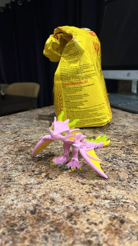Customer's 3D-printed pink and yellow dragon figure sitting on a counter in front of a bag.