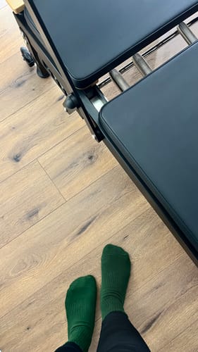 Reviewer wearing alpine green Dual-Direction Grip Basics socks while standing next to a pilates reformer on a wood floor.