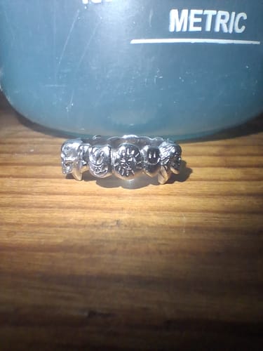 Close-up of a customer's silver-colored Flooded Grill Ring with carved faces, sitting on a wooden surface.