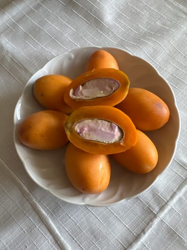 Kundenbild von ganzen und halbierten Mangopflaumen in einer Schale, die das orange Fruchtfleisch und den Kern zeigen.