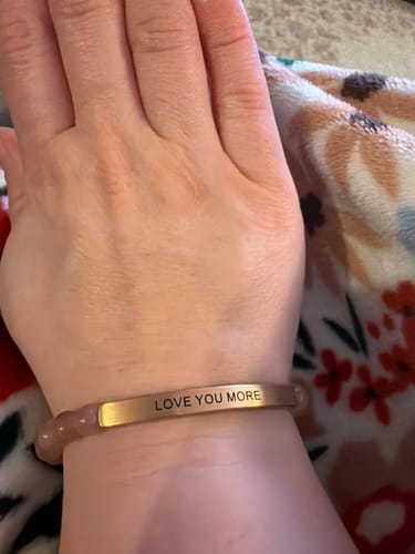 A close-up of a reviewer wearing the LOVE YOU MORE bracelet, showing its rose-gold engraved bar and pink gemstone beads.