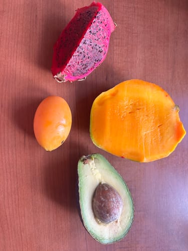Kunde zeigt eine einzelne Marian-Pflaume neben einer aufgeschnittenen Mango, Avocado und Drachenfrucht.