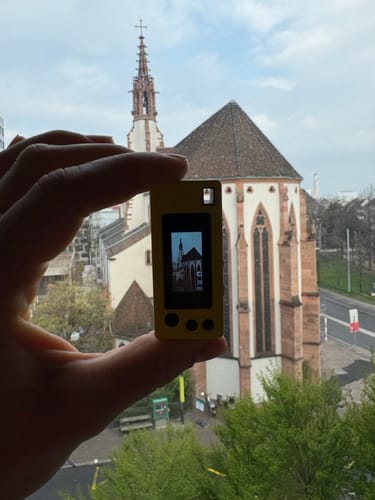Customer holds the yellow RetroShot-1984 camera, with a church visible on its screen and in the background.