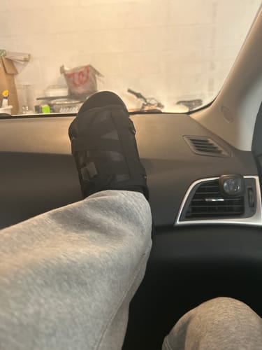 Reviewer wearing the black slides with grey sweatpants, resting their foot on a car dashboard.