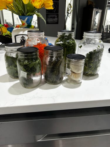 Customer's Chalkboard Top Lids on several glass jars of fresh herbs and vegetables on a kitchen counter.