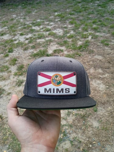 Customer's hand holding a dark gray Florida Plate Hat with a custom license plate patch that reads 'MIMS'.